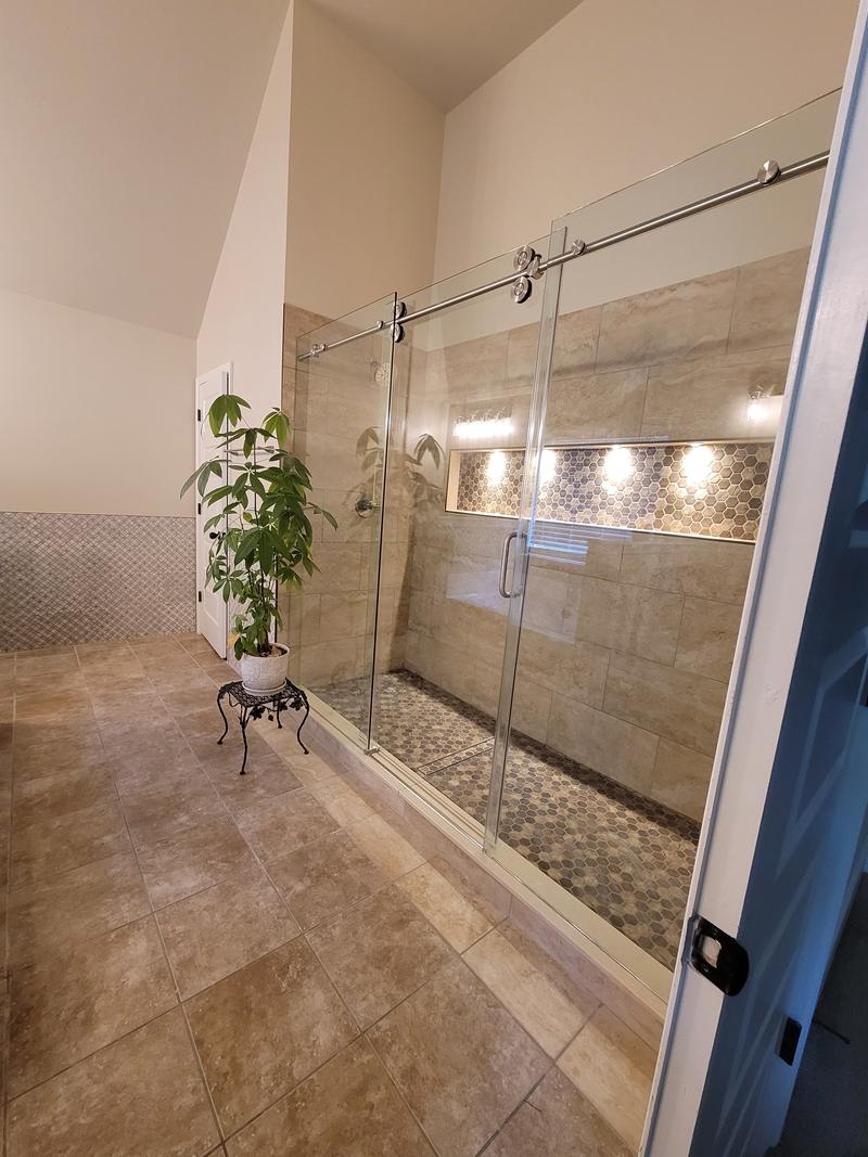 A tiled walk-in shower with a glass door, large-format wall tile, a pebble mosaic floor and an illuminated tiled niche.