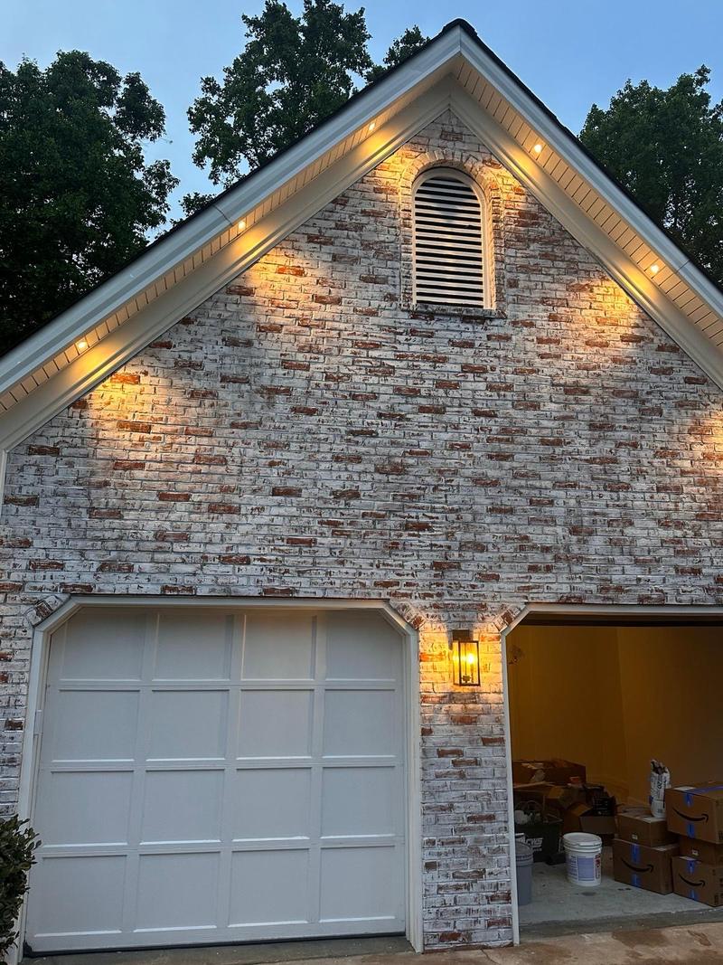 A gable end in mortar-washed white brick lit by soffit lights at dusk, above a white garage door and painted trim.