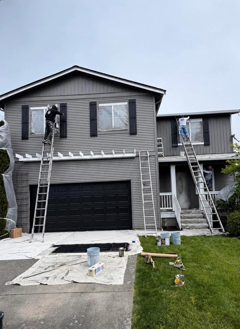 Two painters on ladders coating the siding and trim of a two-story gray house, with drop cloths spread over the driveway and lawn.