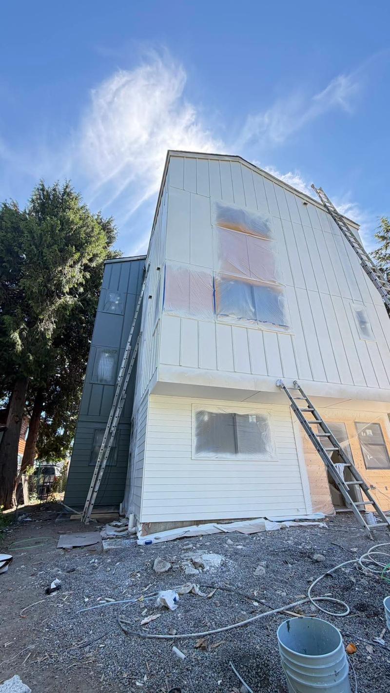 A tall house being spray painted, with the windows masked in plastic and ladders set against the siding.