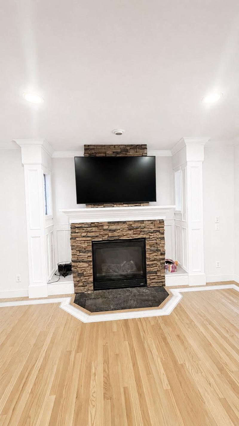 A finished living room with white painted columns and wainscot, a stone fireplace surround and light wood flooring.