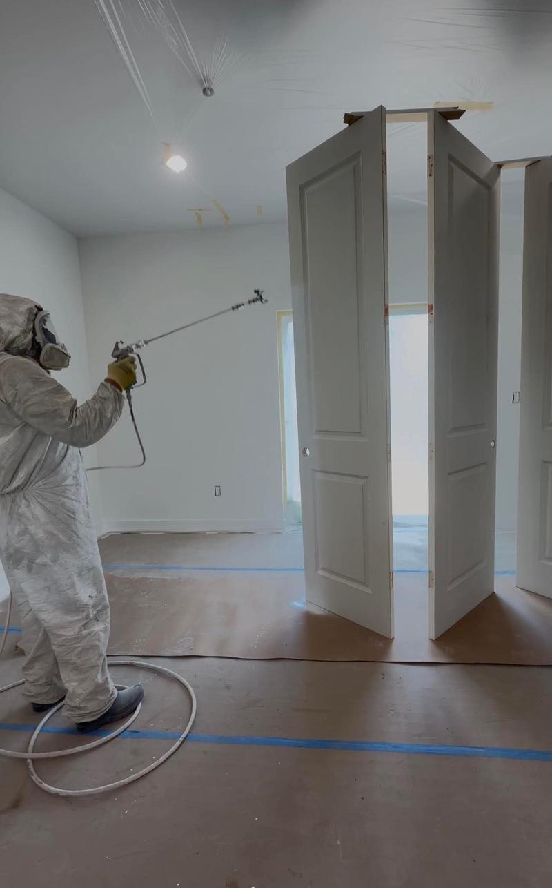 A painter in a protective suit and respirator spray-finishing interior doors, with plastic on the ceiling and paper taped across the floor.
