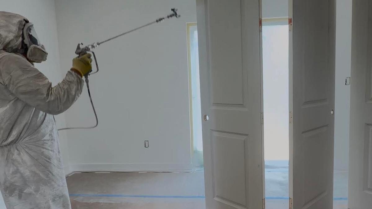 A painter in a protective suit and respirator spray-finishing interior doors, with plastic on the ceiling and paper taped across the floor.