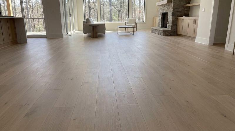 A wide-plank light oak floor running the length of an open living area, toward a stone fireplace and a wall of windows.
