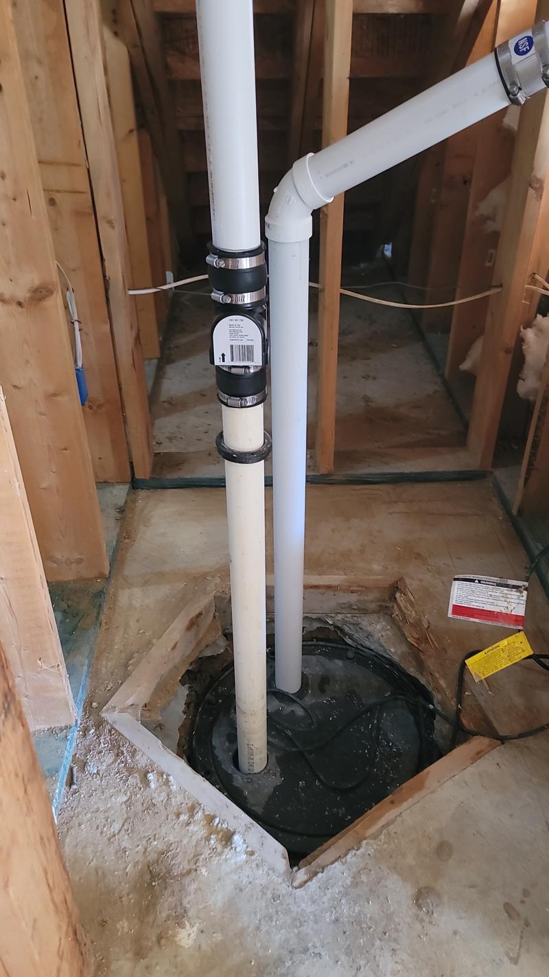 PVC drain standpipes and a check valve rising from a sealed sump basin lid in a framed basement floor.